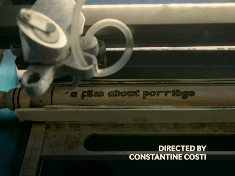 A wooden spurtle sits inside an automated woodburning machine, which has just finished burning "A film about porridge" into the shaft of a spurtle. On the screen simple white tet reads "directed by Constantine Costi"